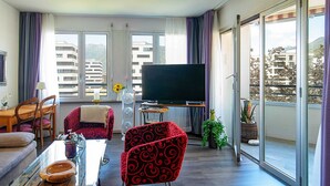Un salon moderne doté d’un téléviseur à écran plat, d’une table à manger avec des chaises, d’un canapé et d’un balcon offrant une vue sur les bâtiments et les montagnes.