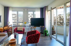 Ein modernes Wohnzimmer mit Flachbildfernseher, Esstisch mit Stühlen, einer Couch und einem Balkon mit Blick auf Gebäude und Berge.