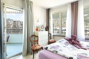 Habitació amb un llit, una cadira, una taula petita, un ventilador i vistes a edificis per la finestra.
