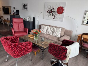 Un salon comprenant un canapé, deux fauteuils rouges à motifs, une table basse avec un vase de fleurs, un fauteuil rouge et un coin repas avec une table et des chaises en bois.