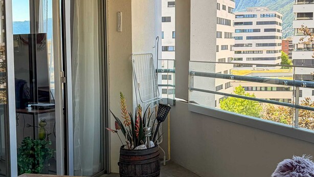 Un balcon avec une table en bois, des chaises en osier, une plante en pot et une vue sur des immeubles et des montagnes.