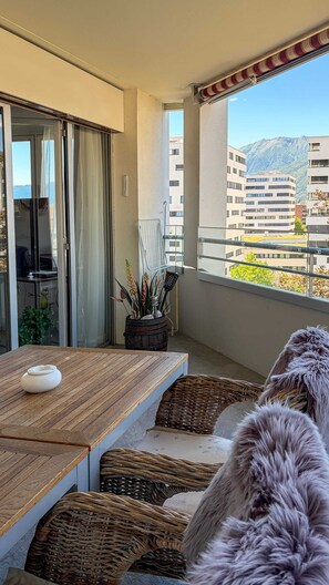 Un balcó amb una taula de fusta, cadires de vimet, una planta en test i vistes a edificis i muntanyes.