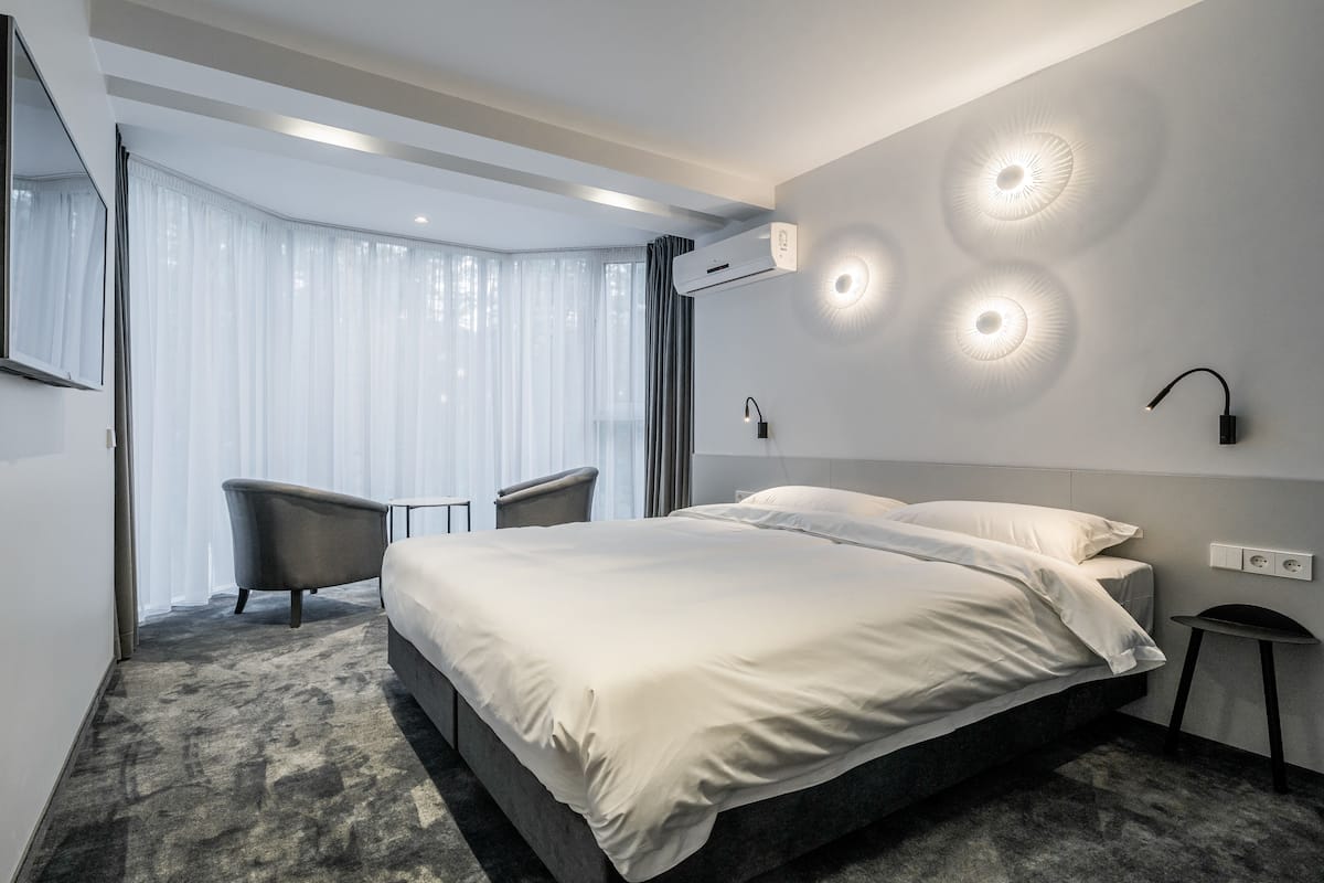 A modern hotel room with a large bed, a seating area with a chair, and a large window with curtains.