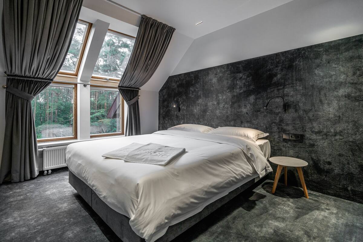A modern bedroom with a large bed, a small round table, and a view of the outdoors through large windows.