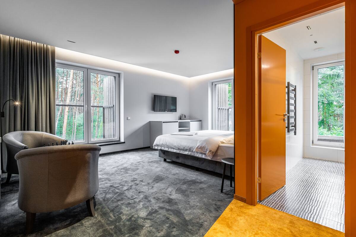 A modern hotel room with a large bed, a seating area with a chair, a TV mounted on the wall, and large windows offering a view of the outdoors.