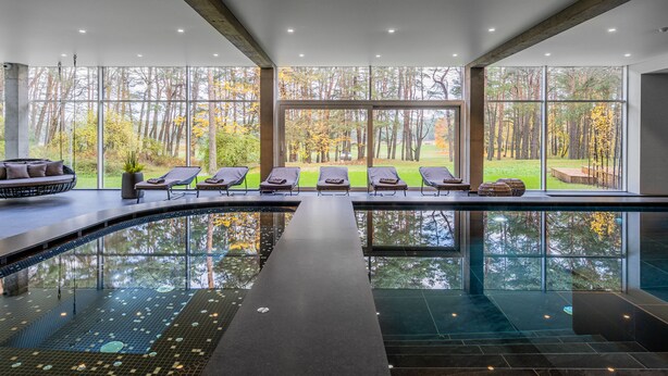An indoor swimming pool with clear glass walls, lounge chairs, and a view of a forested area.