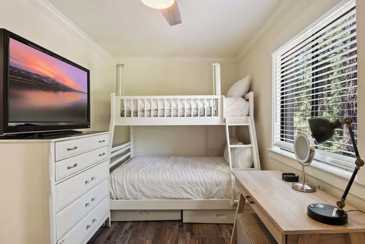 A bunk bed room with a desk, drawers, a TV, and a window with blinds.