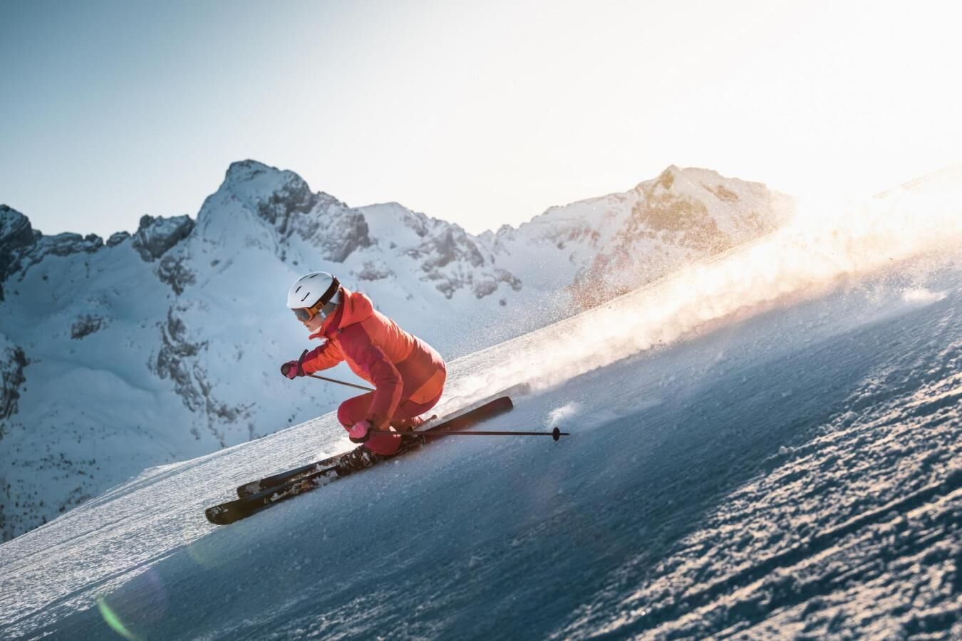Un skieur vêtu d’une combinaison rouge dévale une pente enneigée, avec des montagnes en arrière-plan.