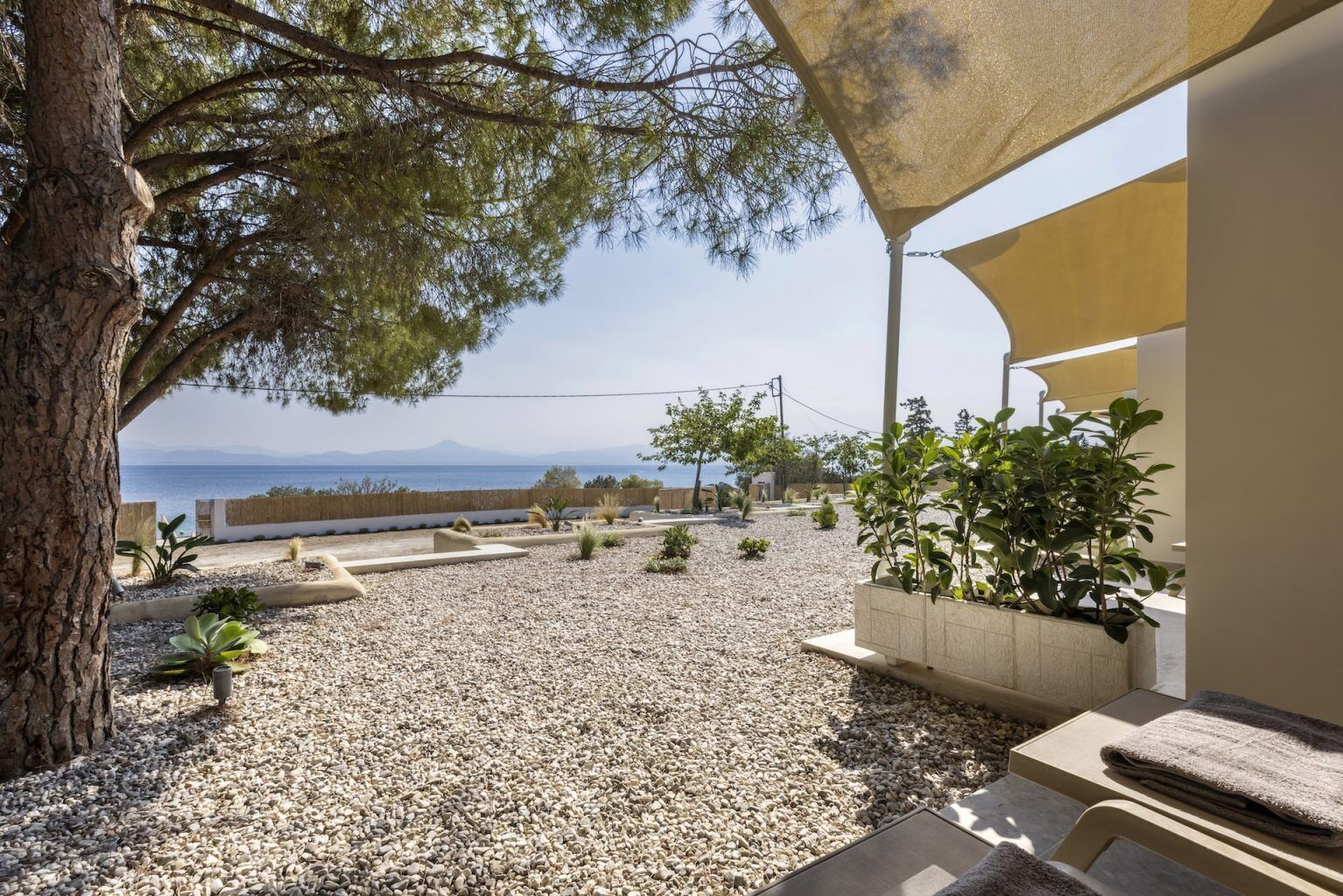 A patio area with a view of the sea, surrounded by gravel, plants, and a pergola.