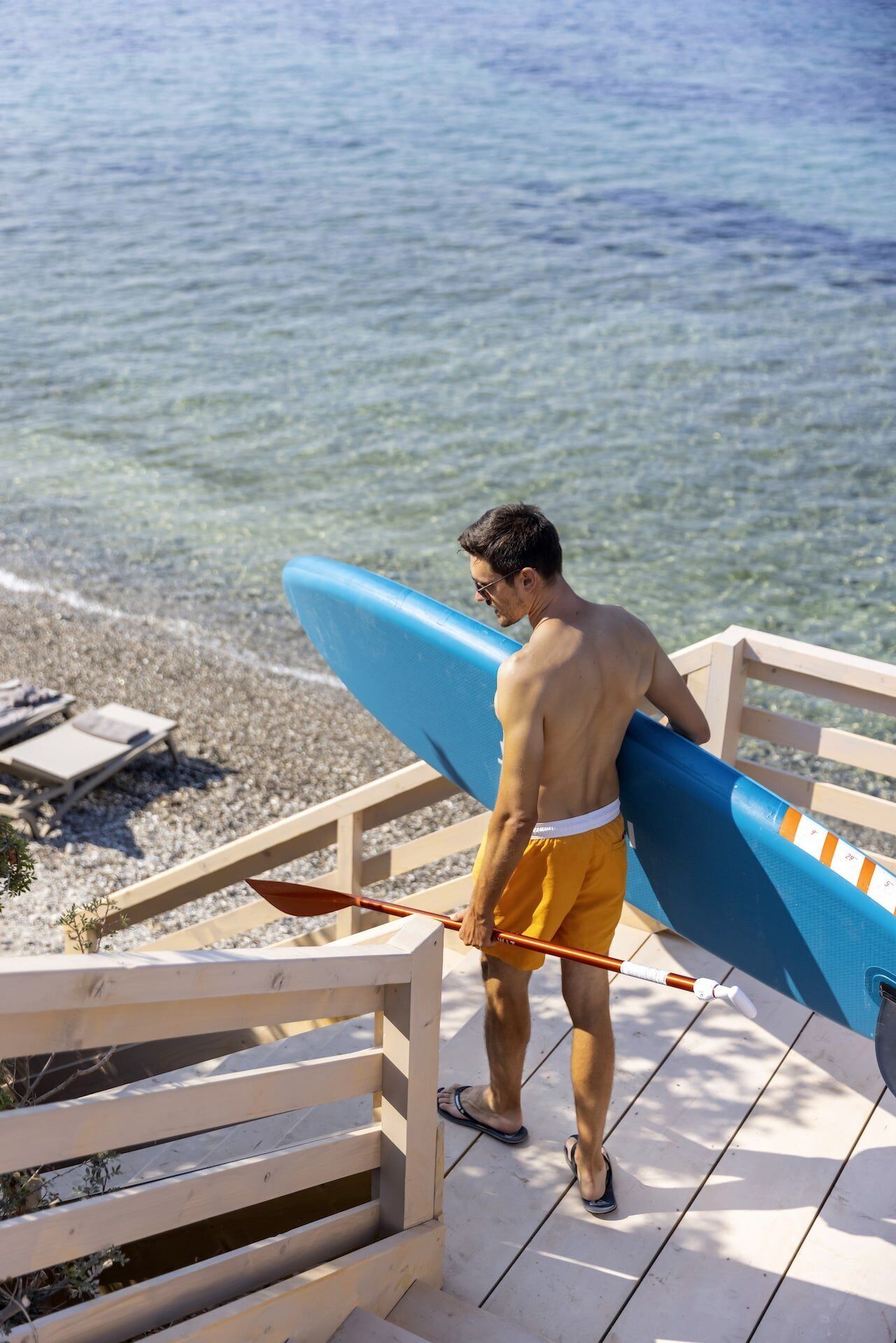 A man carrying a blue surfboard on a wooden deck by a beach.