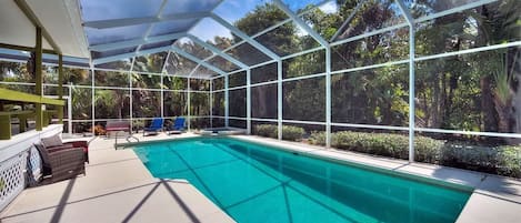 A covered swimming pool with a glass enclosure, surrounded by greenery and a bench area.