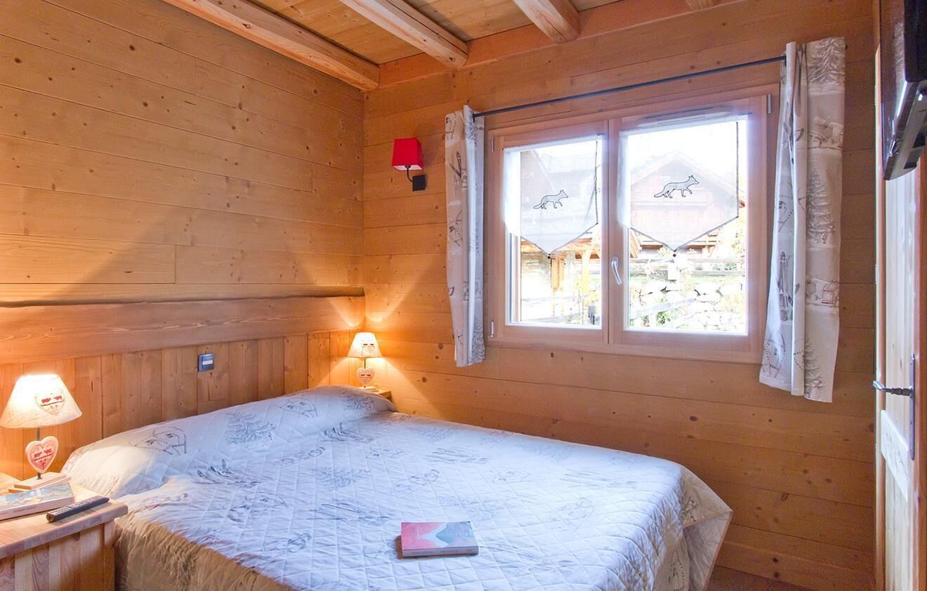 Una habitación de cabaña de madera con una cama, una mesita de noche con lámpara y una ventana con cortinas.