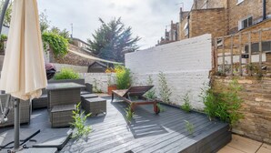 Une terrasse en bois avec des meubles d’extérieur, un mur blanc et un bâtiment en briques en arrière-plan.