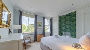 Une chambre avec un grand lit, une table de chevet, une chaise, une fenêtre avec des rideaux, un radiateur et un mur orné d’un motif végétal vert.