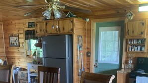 Interior de una cabaña de madera con una mesa de comedor, sillas, un refrigerador, una estufa y un ventilador de techo.