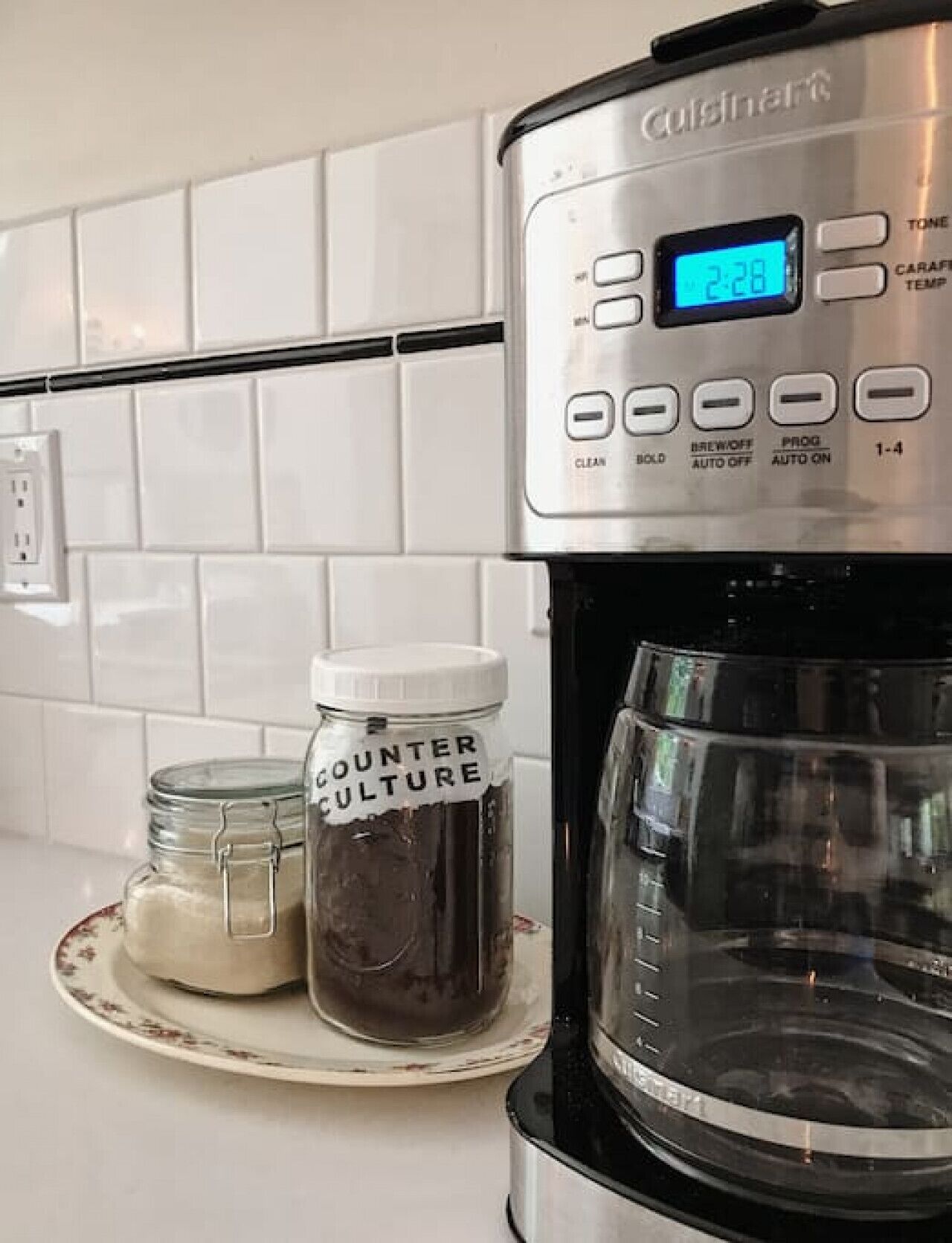Une cuisine avec une machine à café Cuisinart, un bocal étiqueté « COUNTER CULTURE » et une assiette sur laquelle se trouve un petit bocal.