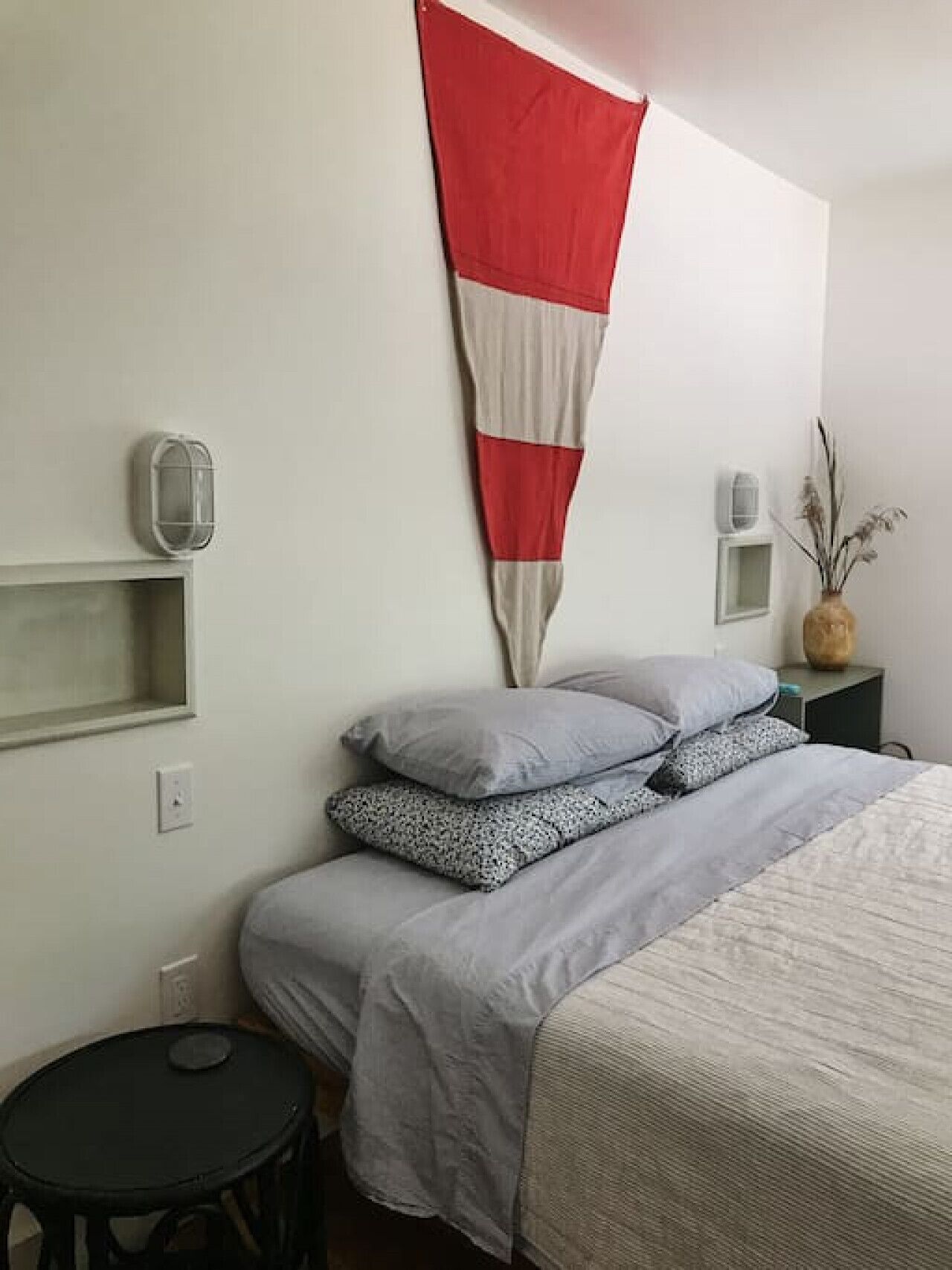 Un dormitorio ordenado con una cama, almohadas, una mesita redonda y un estante en la pared.