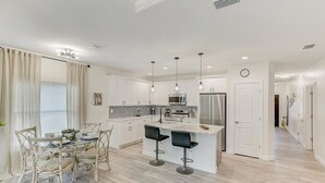 Una cocina moderna con armarios blancos, una isla central con taburetes y luces colgantes, además de un refrigerador de acero inoxidable.