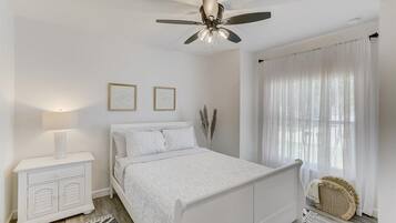 Ein Schlafzimmer mit einem weißen Bett, einem Nachttisch, einem Deckenventilator und einem Fenster mit durchscheinenden Vorhängen.