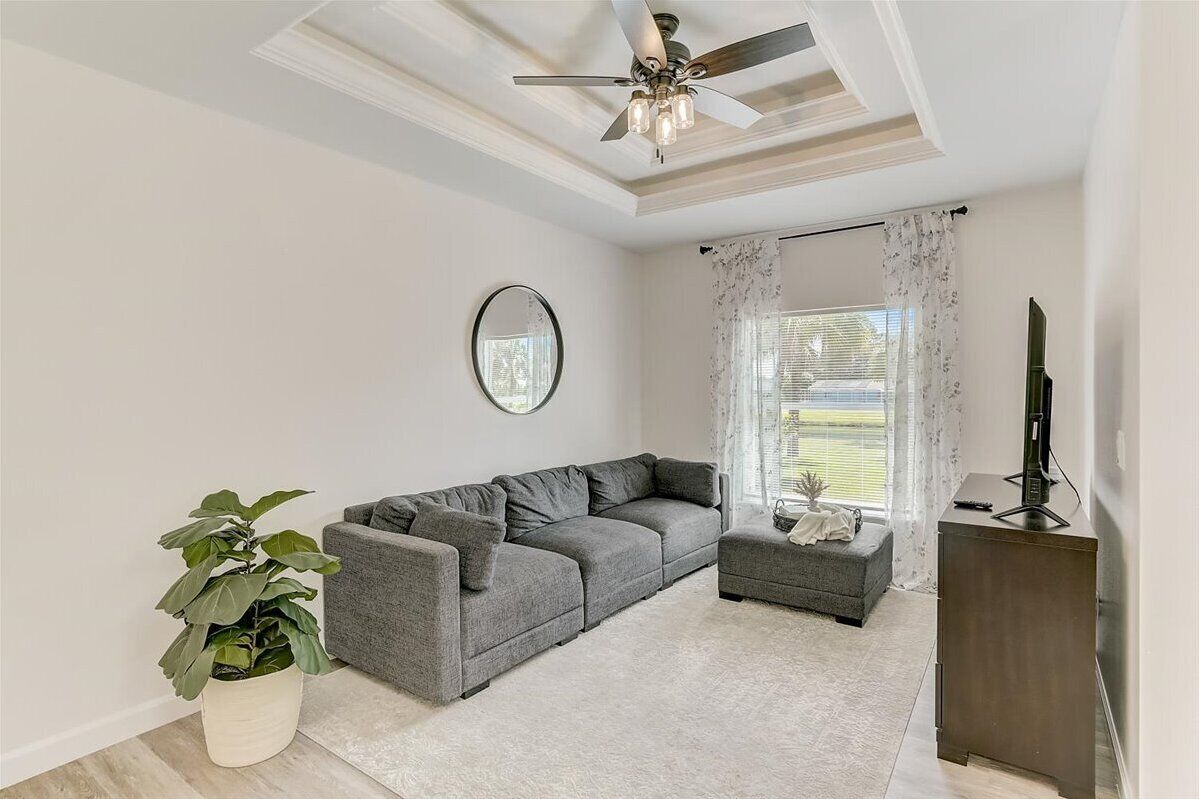 Un salon avec un canapé d’angle gris, un petit pouf, un ventilateur de plafond, un téléviseur à écran plat posé sur un meuble en bois, une plante en pot et une grande fenêtre ornée de rideaux légers.