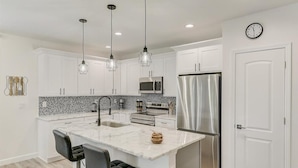 Una cocina moderna con armarios blancos, una isla de mármol, electrodomésticos de acero inoxidable e iluminación colgante.