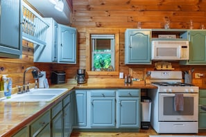 Una cocina con paredes de madera, un lavamanos, armarios, un microondas, una estufa y una ventana con vistas a la vegetación.