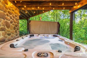 A hot tub with a stone wall backdrop, wooden ceiling, and string lights. - Fern Ridge Cabin - Hot Tub - 60 Acres with Trails! (Laurelville)