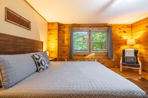 Une pièce aux murs lambrés, avec un lit, une fenêtre équipée de stores, une chaise avec un coussin et un panneau décoratif fixé au mur.