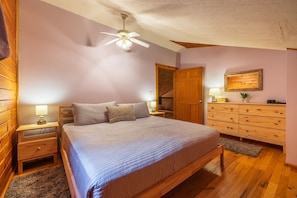 Un dormitorio con cama, mesitas de noche de madera, cómoda, ventilador de techo y una puerta que da a otra habitación.
