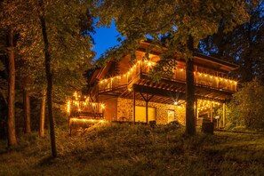 A wooden cabin with a stone facade, surrounded by trees, and illuminated by string lights.