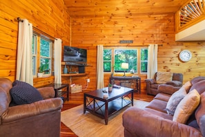 A wooden cabin interior with a TV, leather couches, a coffee table, and a clock on the wall.