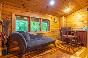 A wooden cabin interior with a sofa, a chair, a desk, and a window with a view of greenery.