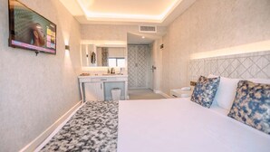 Habitación de hotel con cama, escritorio con espejo, televisión y baño visible a través de una puerta abierta.