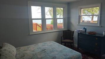 A bedroom with a bed, a chair, a dresser, and two windows with a view of the outdoors.