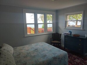 Ein Schlafzimmer mit einem Bett, einem Sessel, einer Kommode und zwei Fenstern mit Blick ins Freie.