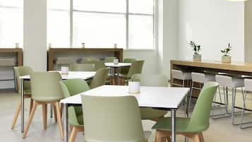 A modern dining area with green chairs and white tables, large windows, and pendant lighting.