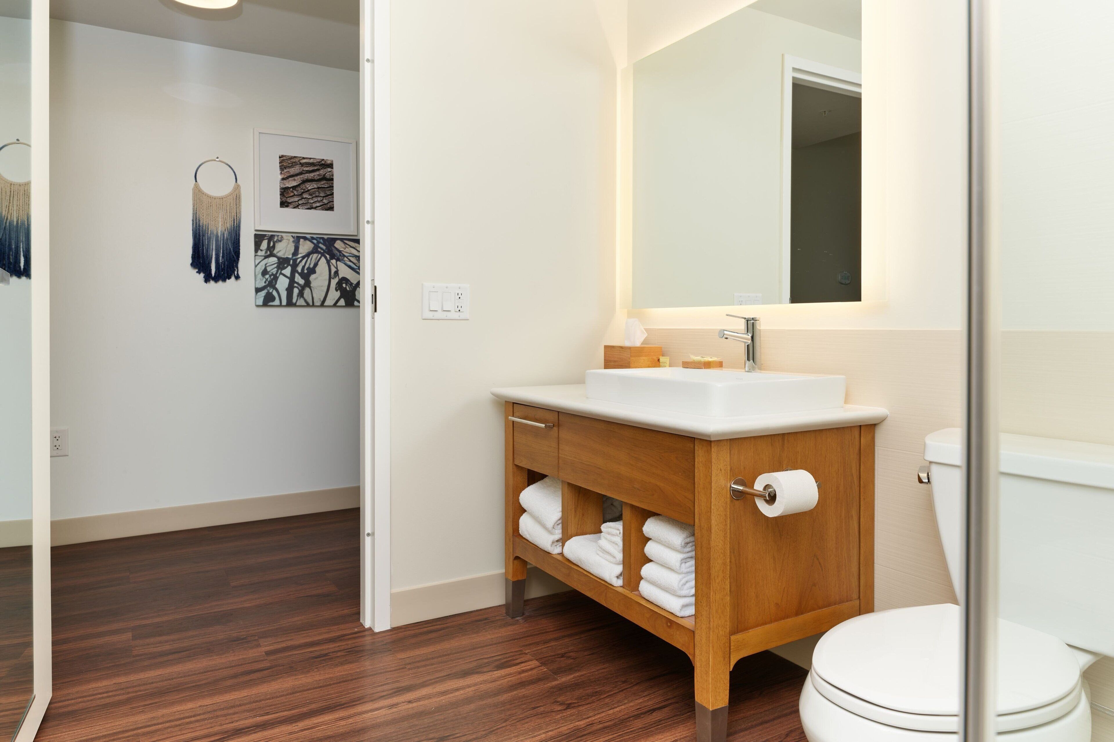 Un baño moderno con tocador de madera, lavamanos blanco y retrete. Hay toallas sobre el tocador y una obra de arte en la pared.
