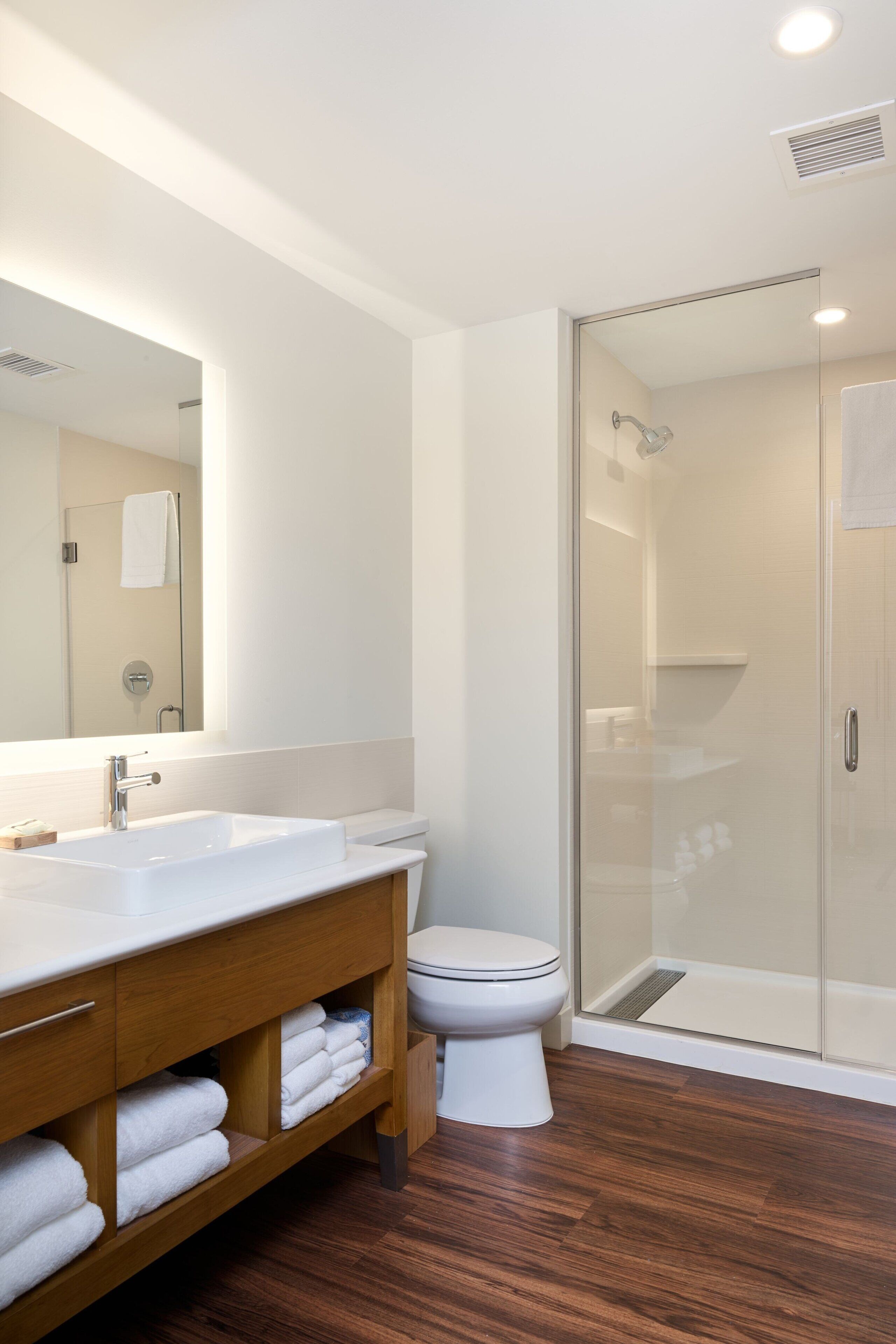 Un baño moderno con ducha envuelta en vidrio, un lavabo blanco y un mueble de tocador de madera con toallas.