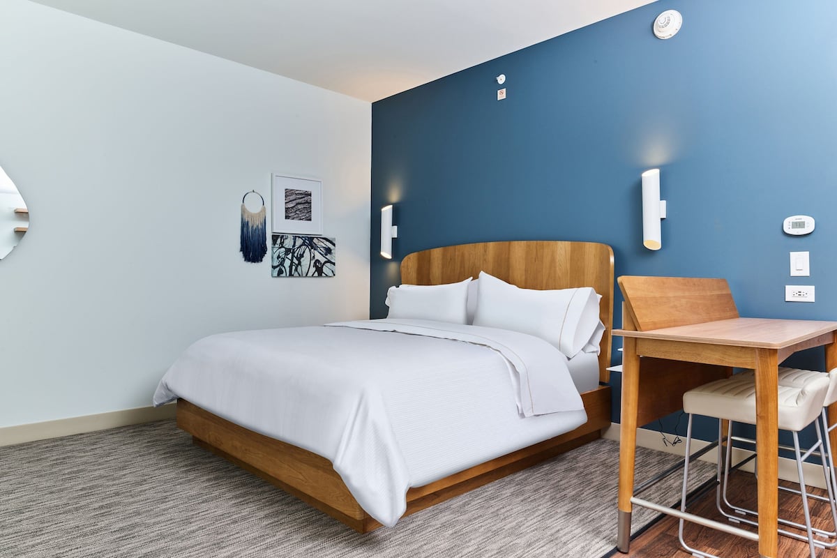 A modern hotel room with a wooden bed, a small desk, and a blue accent wall.