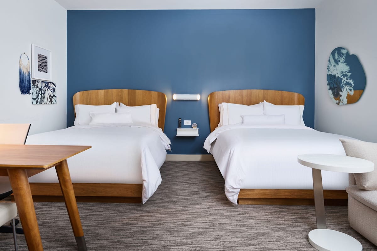 A hotel room with two beds, a small table, a round side table, and a wall-mounted light fixture.