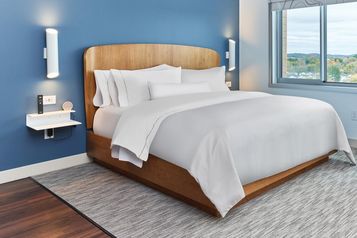 A modern bedroom with a wooden headboard, white bedding, and a view of a tree-lined landscape through a large window.