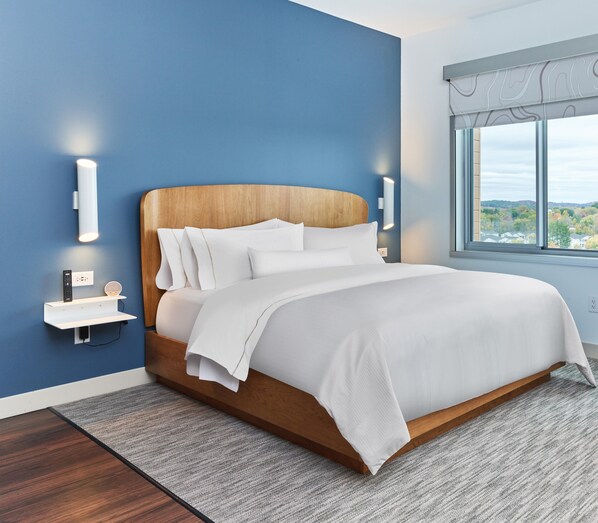 Une chambre moderne avec une tête de lit en bois, une literie blanche et une vue sur un paysage arboré, visible à travers une grande fenêtre.