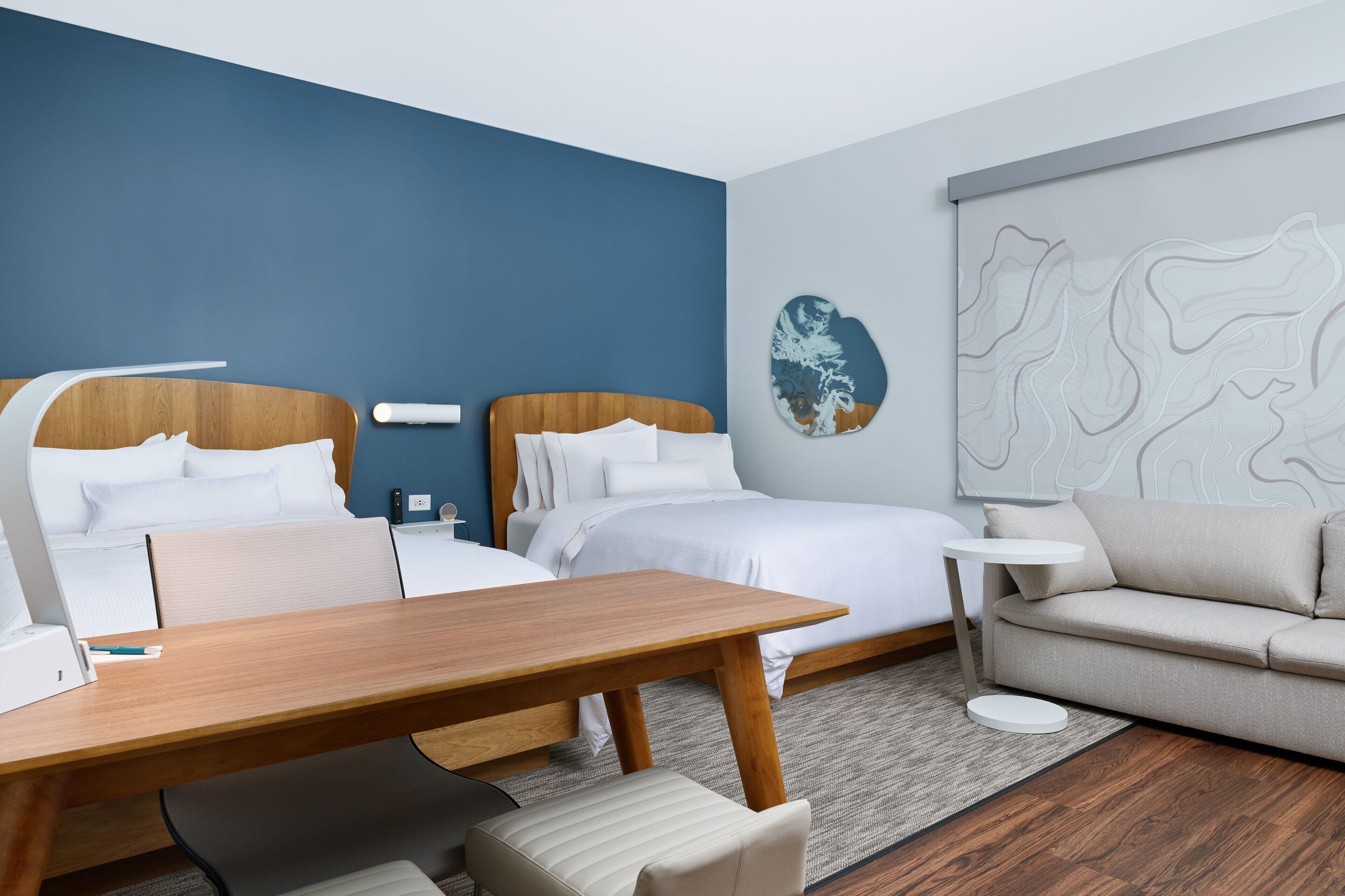 Quarto de hotel moderno com duas camas, uma escrivaninha de madeira, uma mesa de centro redonda, um sofá e uma grande obra de arte abstrata na parede.
