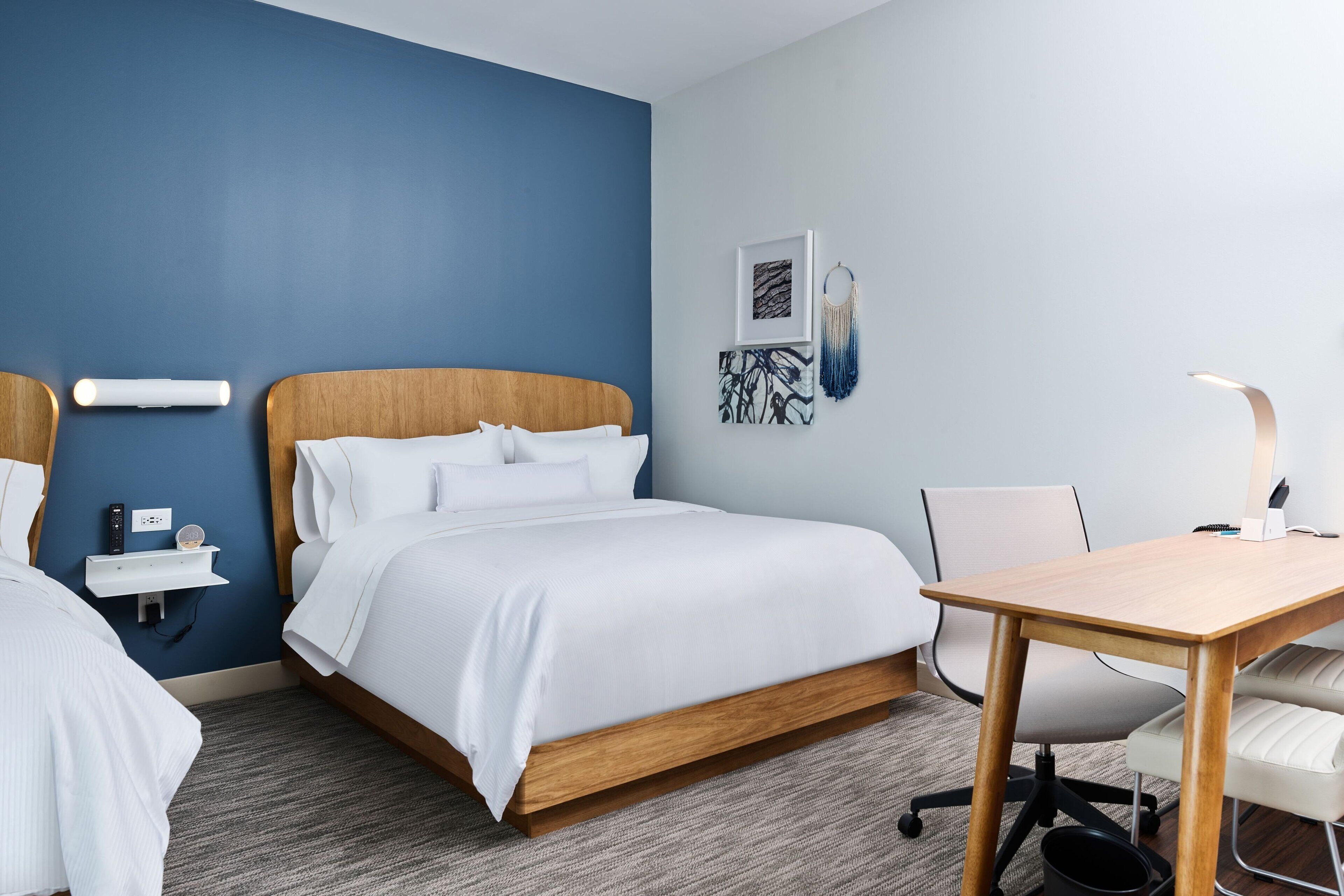 Une chambre d’hôtel avec un grand lit, un bureau et une chaise. Il y a une étagère fixée au mur, sur laquelle se trouvent une télécommande et un téléphone.