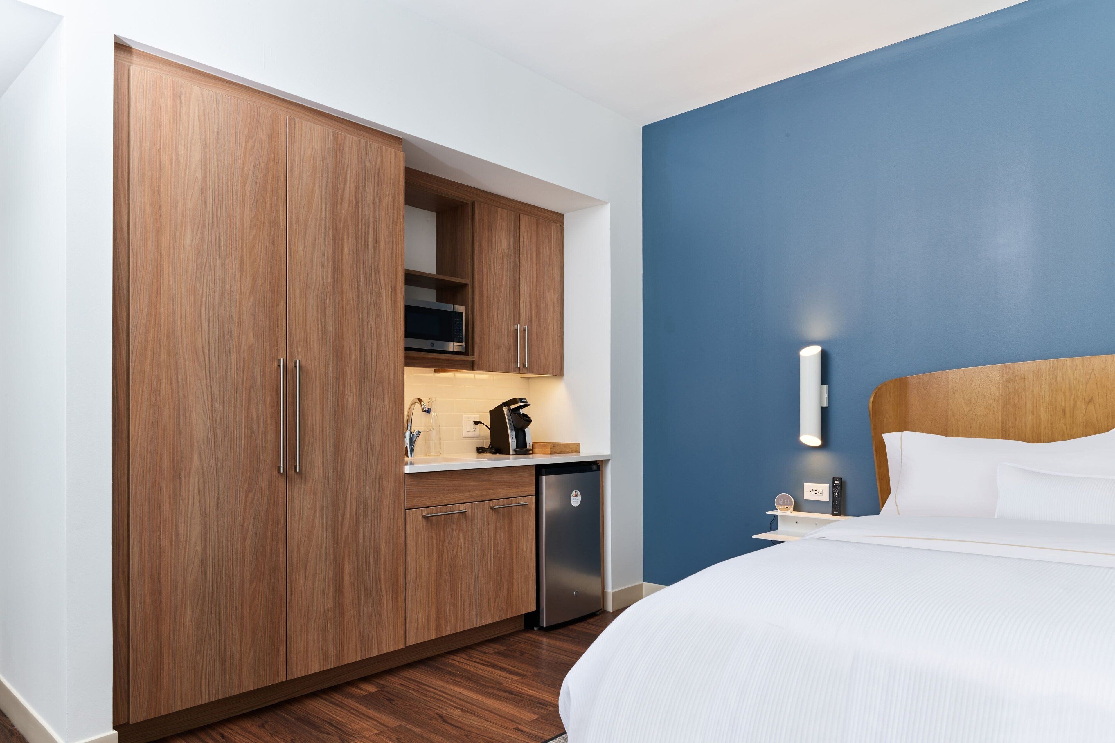 A hotel room with a bed, a wooden wardrobe, a kitchenette with a microwave, and a blue accent wall.