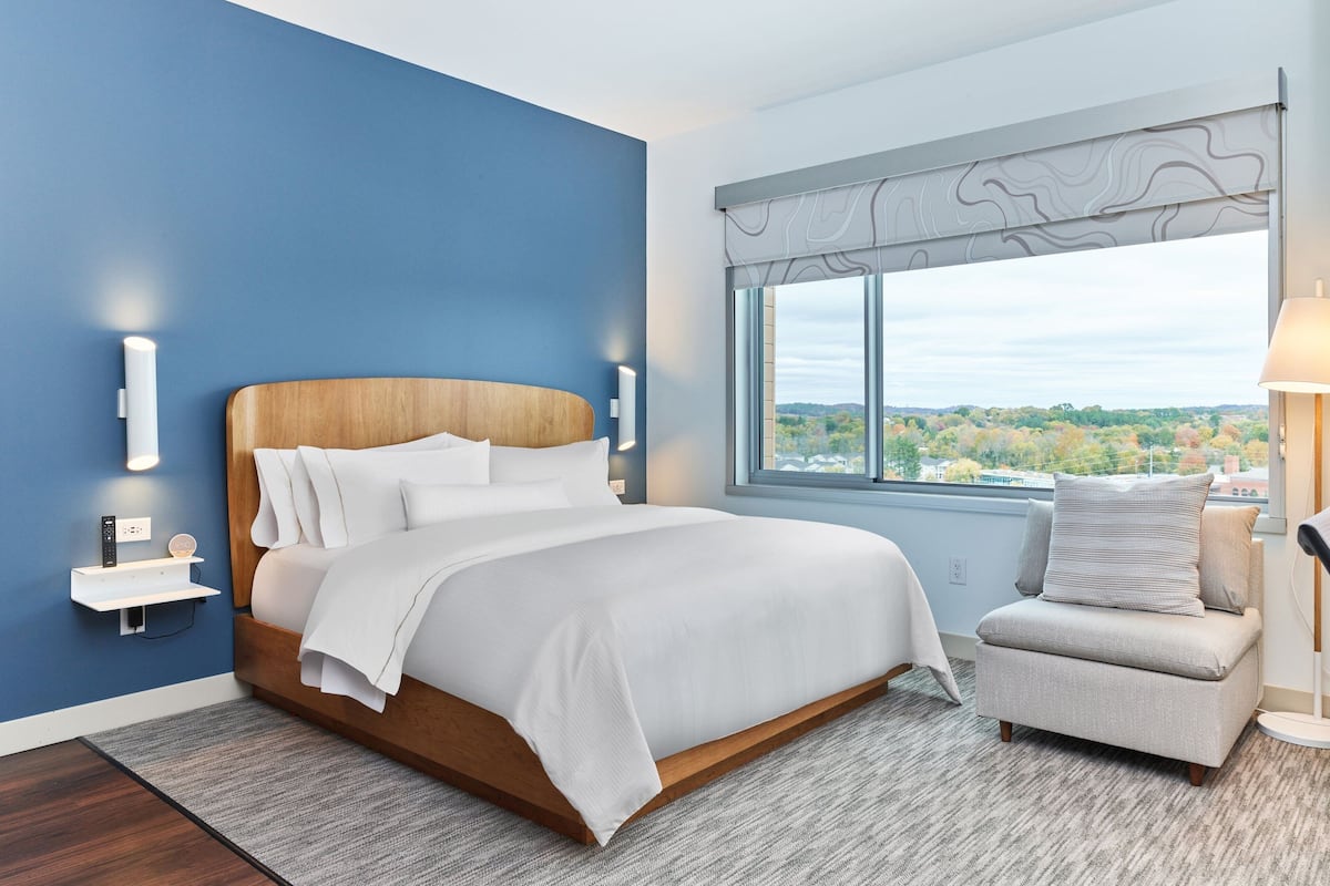 A modern bedroom with a large bed, a nightstand, a chair, and a window with a view.