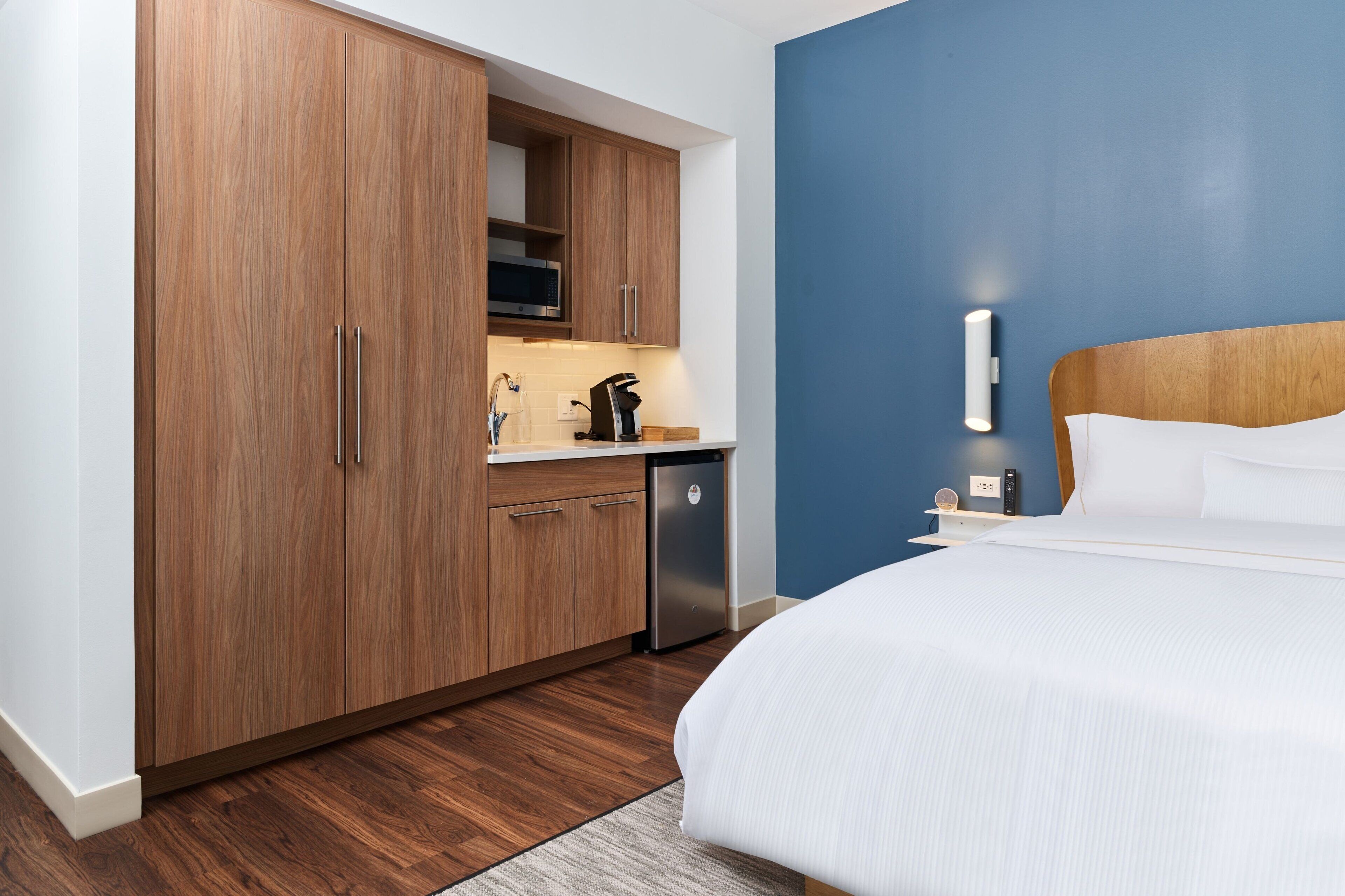 A hotel room with a wooden wardrobe, a kitchen area with a microwave and coffee machine, a bed with white bedding, and a blue accent wall.