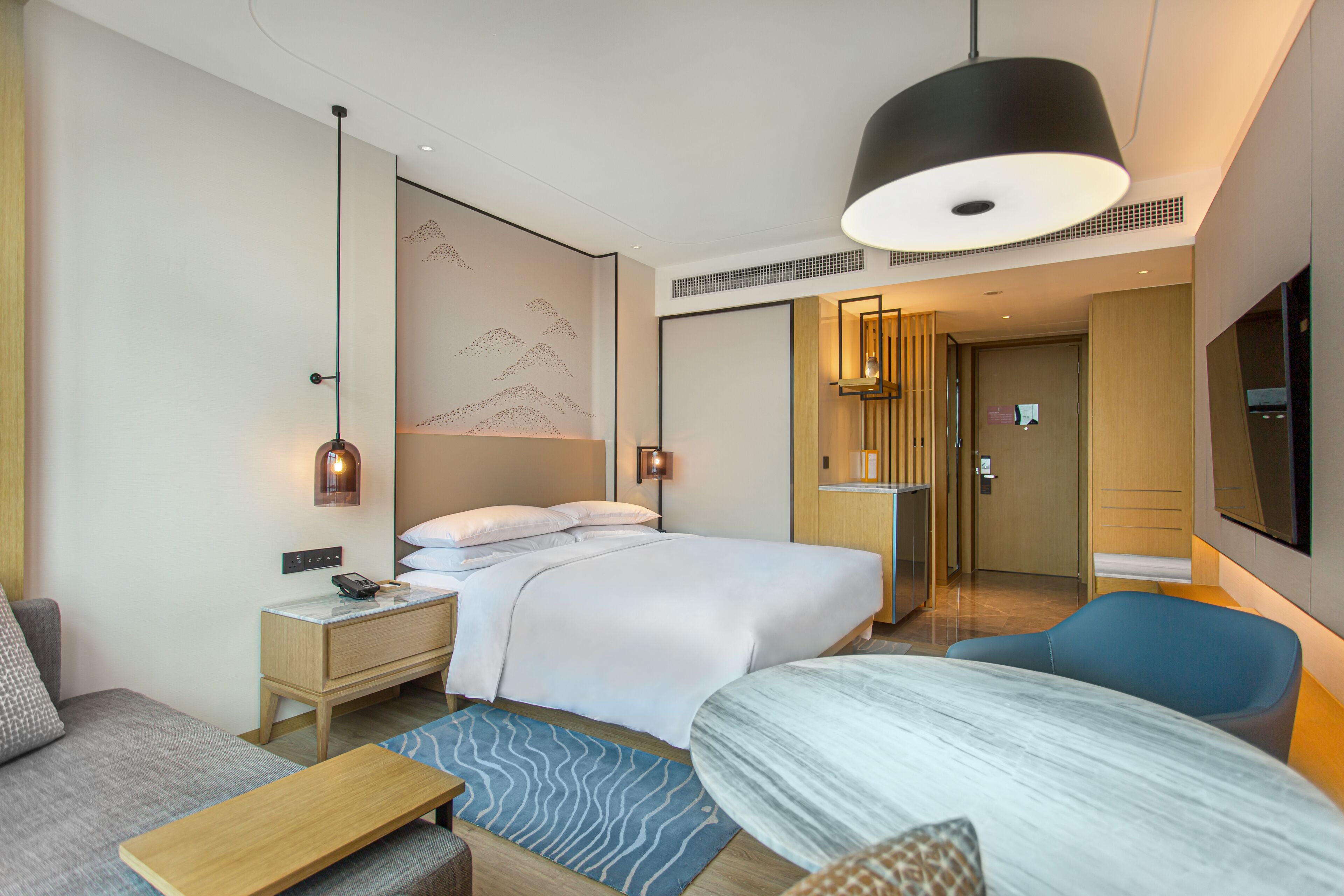 Une chambre d’hôtel moderne équipée d’un lit, d’un bureau, d’une chaise, d’une télévision et d’une salle de bain visible à travers une porte ouverte.