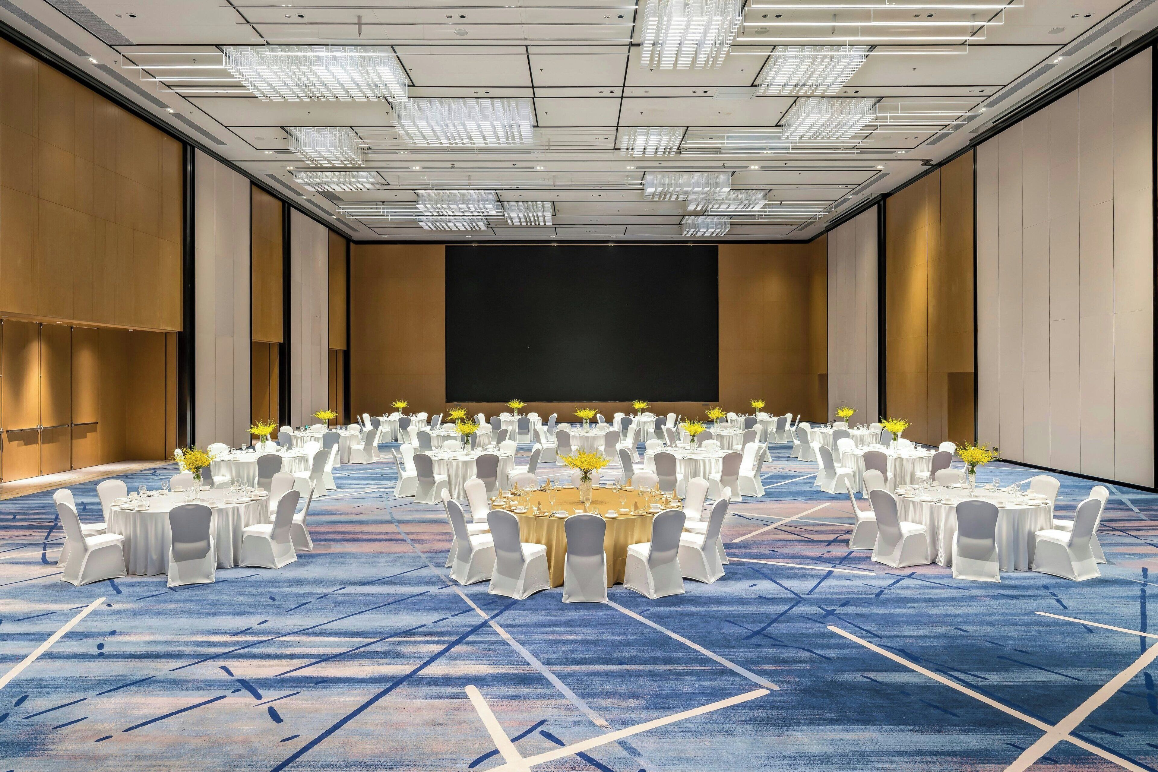 A conference room with a large screen, multiple round tables with chairs, and a carpeted floor.