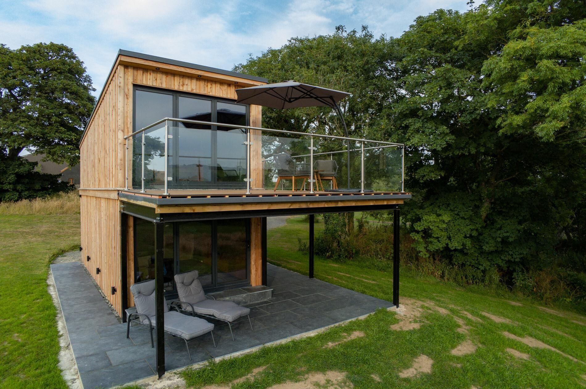 A modern wooden house with a glass balcony, outdoor seating, and a view of the surrounding greenery.